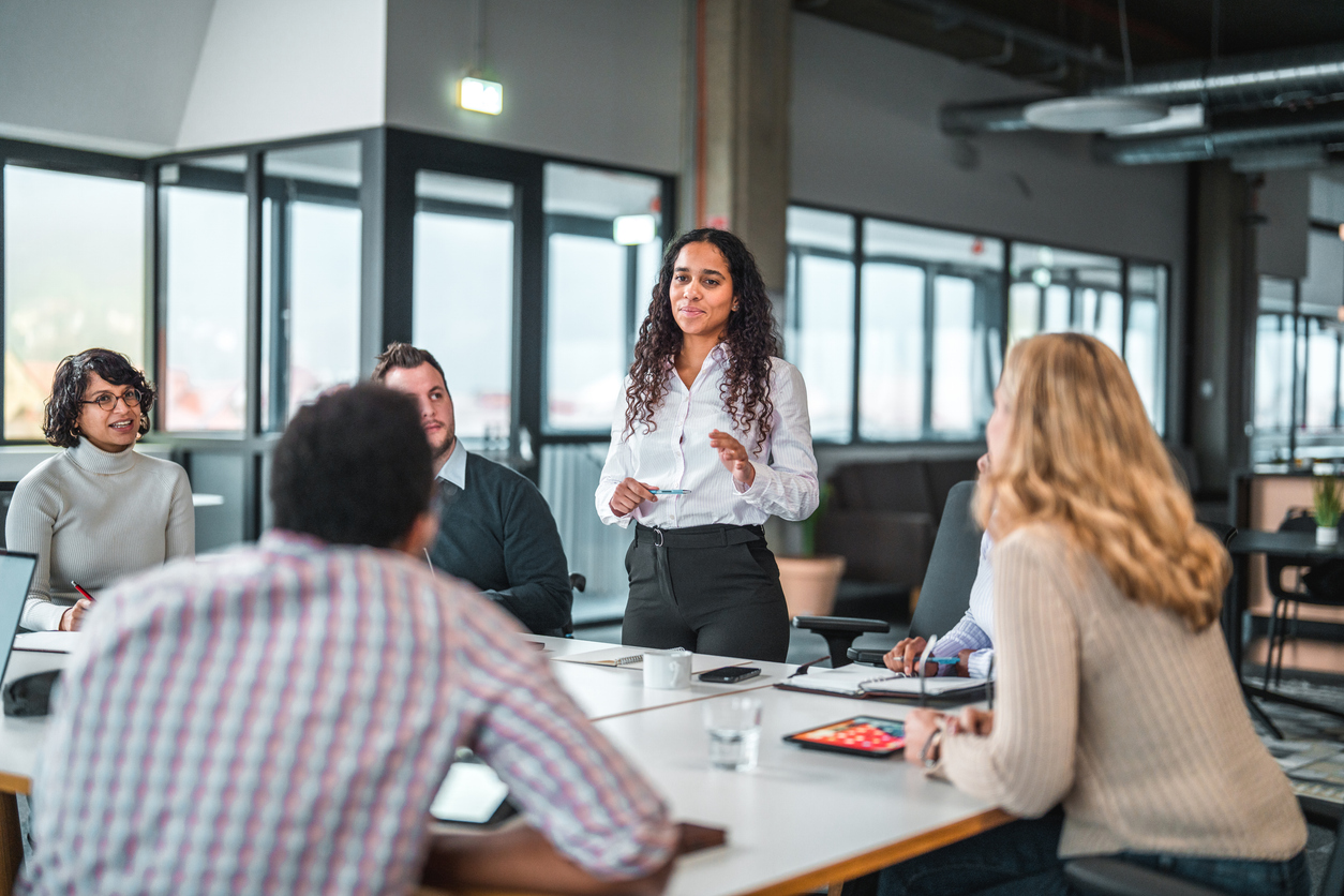 A Manager Stands While Addressing a Team of Colleagues Gathered Around a Conference Table